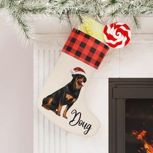 May include: A personalized Christmas stocking with a red and black buffalo plaid cuff, a white background, and a Rottweiler dog wearing a Santa hat. The stocking is personalized with the name "Doug".