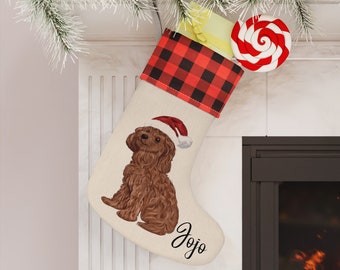 Op de afbeelding: Een rode en zwarte kerstsok met buffalo plaid met een bruine poedel die een kerstmanmuts draagt en de naam "Jojo" geborduurd op de sok.