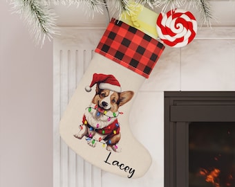 Op de afbeelding: Een gepersonaliseerde kerstsok met een rode en zwarte buffalo plaid manchet. De sok heeft een schattige illustratie van een corgi-hond die een kerstmanmuts draagt en is ingepakt in kerstverlichting. De naam "Lacey" is geborduurd op de sok.