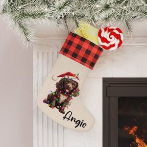 May include: A personalized Christmas stocking with a brown and white dog wearing a Santa hat and surrounded by Christmas lights. The stocking is red and black plaid with a white background and the name "Angie" is written in black cursive.