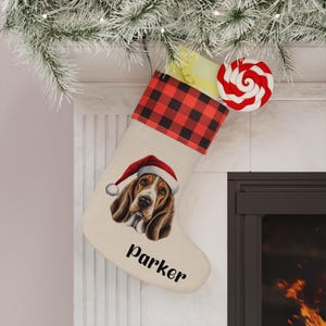 Puede incluir: Un calcetín navideño con un Basset Hound con un gorro de Papá Noel. El calcetín tiene un puño a cuadros rojos y negros y el nombre "Parker" impreso en la parte delantera. Una piruleta roja y blanca está adjunta.