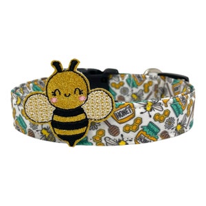 May include: A white dog collar with a black and gold bee charm. The collar has a white background with a repeating pattern of honey pots, honeycombs, and bees. The text "Honey" is printed on the collar.
