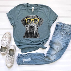 Pode incluir: Uma camiseta azul turquesa com uma ilustra&ccedil;&atilde;o em preto e branco de um c&atilde;o usando &oacute;culos de sol amarelos. O c&atilde;o est&aacute; olhando diretamente para a c&acirc;mera. A camiseta &eacute; combinada com jeans azul claro e t&ecirc;nis brancos.