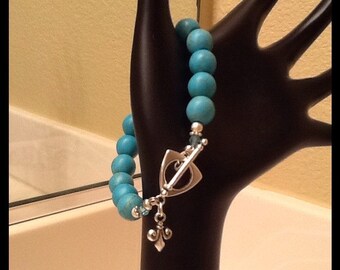 May include: A turquoise beaded bracelet with a silver clasp and a decorative charm. The bracelet is displayed on a black hand.