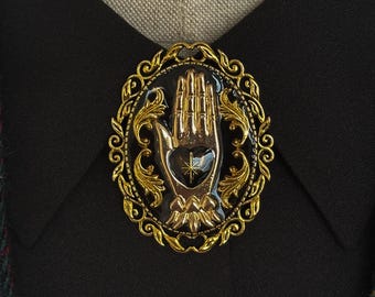 May include: A gold-coloured brooch with a hand design inside an oval frame. The hand is raised, with detailed fingers. The brooch has a black background and is set against a black shirt.