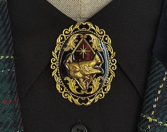 May include: An oval brooch featuring a gold-toned filigree frame surrounding a dark centre. The centre displays a raised gold bird figure perched on a branch against a deep red diamond-shaped background with a small gold star symbol above the bird.
