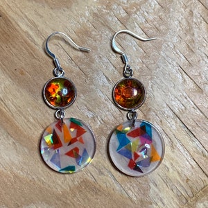 Puede incluir: Un par de pendientes de aro de plata con una piedra redonda, iridiscente, naranja y amarilla y un dije redondo de resina transparente con confeti de colores.