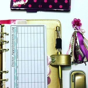 May include: A gold-colored planner with a white page open to a doctor's appointment log. The log has columns for date, reason, tests, results/diagnosis, instructions, and medication. A gold pen with glitter inside is clipped to the planner.
