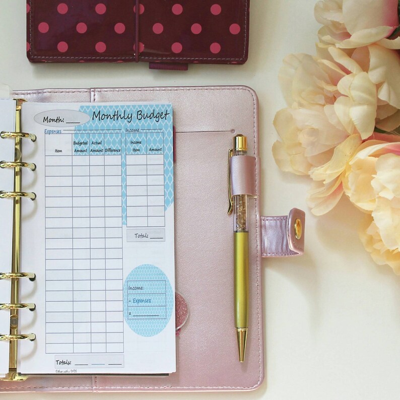 May include: A pink leather-bound planner with a monthly budget tracker insert. The planner has a gold ring binder and a pen with glitter inside is tucked into the side pocket.
