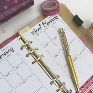 May include: A gold binder with a white meal planning page open. The page has a grid with days of the week labeled. A gold pen with glitter is lying on the page.