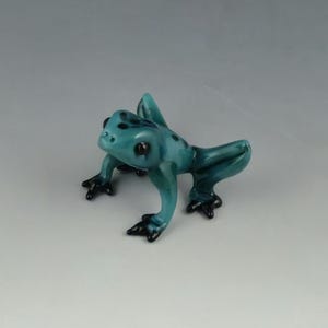May include: A teal-colored glass frog figurine with black spots and feet. The frog is in a crouching position, with its front legs extended and its back legs bent. The figurine is a decorative item.
