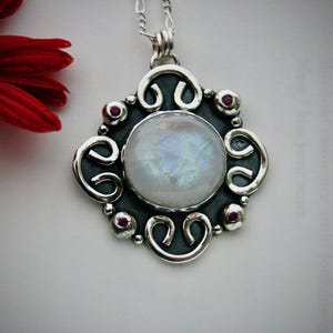 May include: A silver pendant with a large, white moonstone surrounded by a silver filigree design. The pendant has six small, red gemstones around the moonstone.
