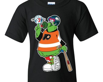 May include: Black t-shirt featuring a cartoon mascot wearing a Philadelphia Flyers jersey and holding a baseball bat. The mascot is green with a red nose and a white hat with a Flyers logo.