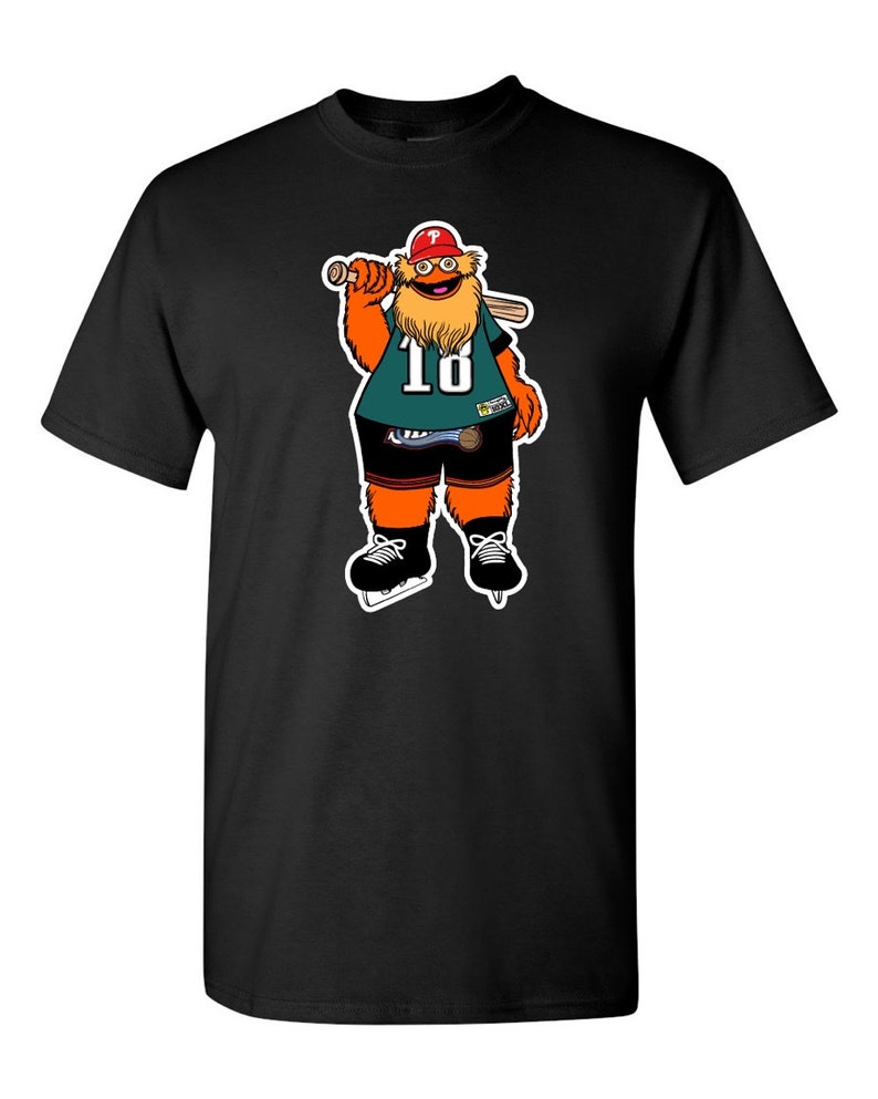 May include: Black t-shirt with a cartoon mascot wearing a green jersey with the number 18, a red hat, and holding a baseball bat. The mascot has an orange body, a brown beard, and is wearing black ice skates.