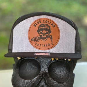 May include: Black and gray trucker hat with a brown leather patch that says "Blue Collar Bastard" and features a skull wearing a hard hat and holding a wrench in its teeth.