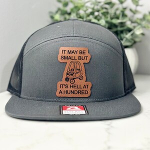 May include: A gray and black trucker hat with a brown leather patch that says "It may be small but it's hell at a hundred" with a cartoon image of a small car.