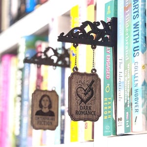 May include: Wooden bookshelf decor featuring a black filigree design with hanging wooden plaques. One plaque reads "DARK ROMANCE" with a heart and sword graphic. Another reads "CONTEMPORARY FICTION". Books are visible in the background.