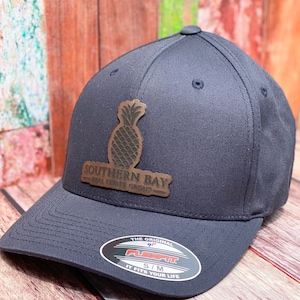 May include: A dark blue baseball cap with a brown leather pineapple patch that says "Southern Bay Real Estate Group". The cap has a Flexfit label and says "S/M It Fits Your Life".