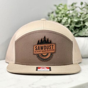 May include: A tan and brown trucker hat with a brown leather patch that says "Sawdust is just man glitter" with a small image of a saw blade and pine trees.