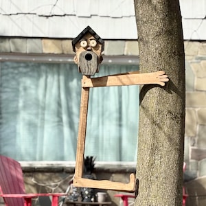 May include: A whimsical wooden birdhouse shaped like a face with a beard and a small roof. The birdhouse is attached to a wooden frame that appears to hug a tree. The birdhouse is painted in natural wood tones and is designed to attract birds.
