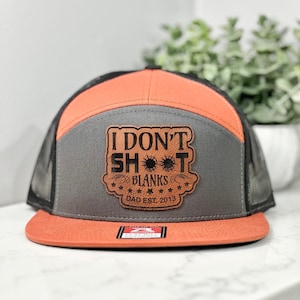May include: A gray and orange trucker hat with a brown leather patch that says "I Don't Shoot Blanks Dad Est. 2013".