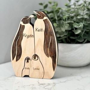 May include: Wooden penguin family sculpture with engraved names: Krystin, Keith, Brielle, and Lorelai. The penguins have dark brown accents and are set against a light wood background. A decorative item for home decor.