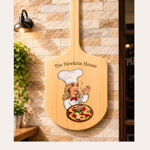 May include: A wooden pizza peel sign featuring the text The Hawkins House, an illustration of a chef in a white hat and uniform making a hand gesture, and a pepperoni pizza graphic. The sign has a light wood grain finish and a long handle.