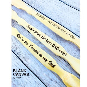 May include: Three wooden back scratchers with engraved text. The text on the scratchers reads "Daddy ~ we got your back!", "Hands down the best DAD ever!", and "You're the Scratch to my Itch". The brand name "BLANK CANVAS by Potter" is also visible.