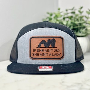 May include: A black and gray trucker hat with a brown leather patch that says "If she ain't 280 she ain't a lady". The patch features a silhouette of a woman in a suggestive pose.