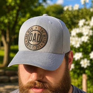 May include: A light gray baseball cap featuring a circular brown patch on the front. The patch displays the text DAD in the center, surrounded by the words THE MAN, THE MYTH, THE LEGEND in a circular border. The cap has a structured crown.