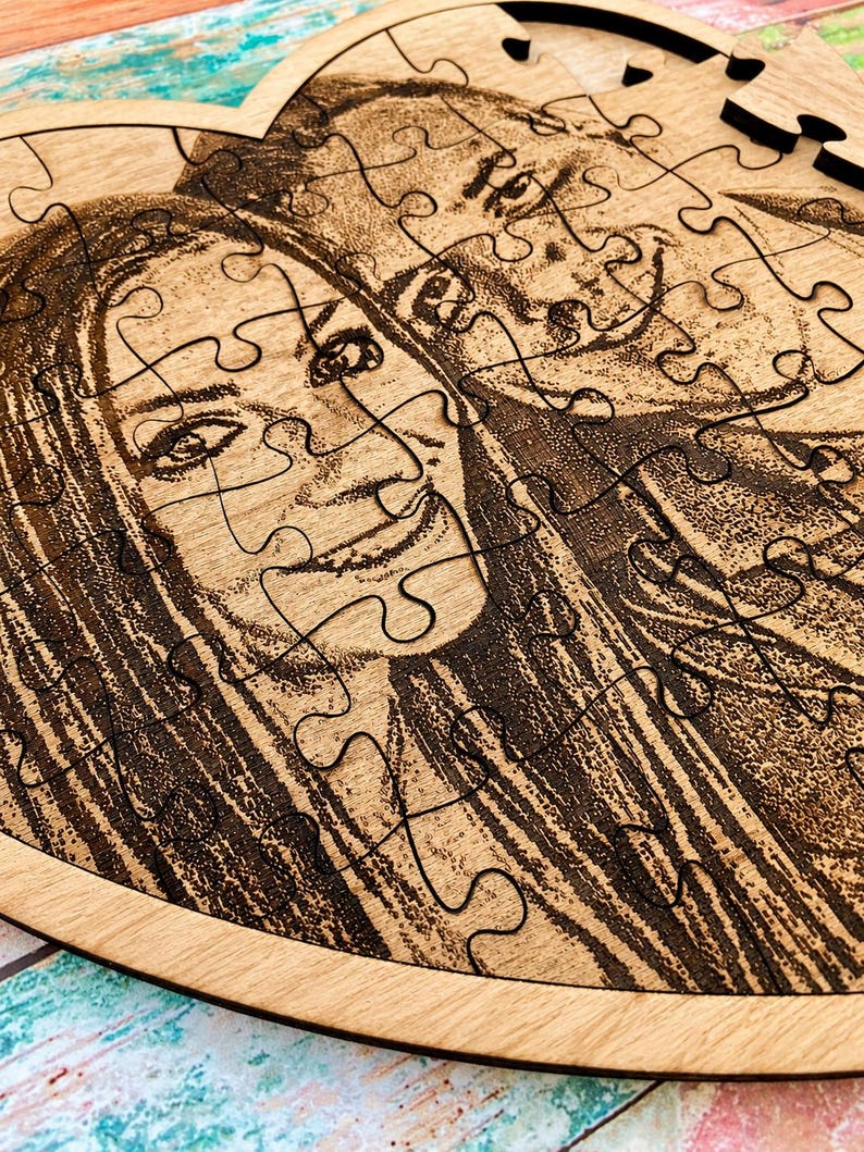 May include: A heart-shaped wooden jigsaw puzzle featuring a laser-engraved portrait of two individuals. The light-colored wood grain serves as the background for the dark, detailed etched image, which is divided into multiple interlocking puzzle pieces.