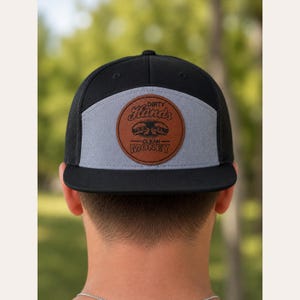 May include: A black and grey trucker hat featuring a circular brown leather patch on the front panel. The patch displays the text Dirty Hands Clean Money with a graphic of two fists. The hat has a flat brim and a mesh back for ventilation.