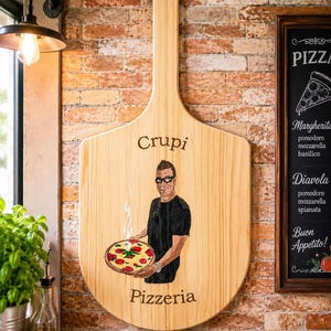 May include: A large wooden pizza peel featuring a hand-painted illustration of a person holding a steaming pizza. The text "Crupi" is printed above the figure, and "Pizzeria" is printed below. The wood grain is visible throughout the surface.