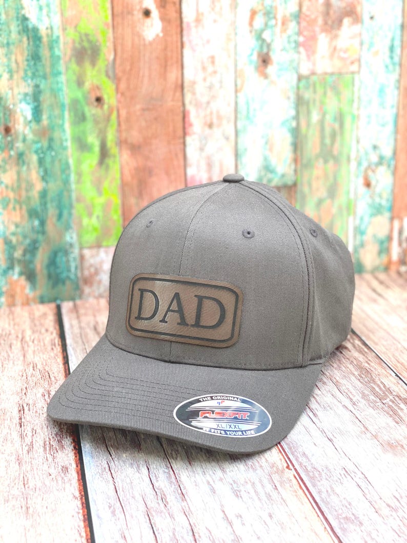 May include: A grey baseball cap with a brown leather patch displaying the word "DAD". The cap features a curved peak and a fitted design. The hat is an XL/XXL size.