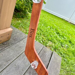 Puede incluir: Un palo de hockey de madera con dos vasos de chupito de vidrio transparente unidos a &eacute;l. El palo est&aacute; te&ntilde;ido de un marr&oacute;n rojizo oscuro y tiene la palabra "BOX" grabada en &eacute;l.