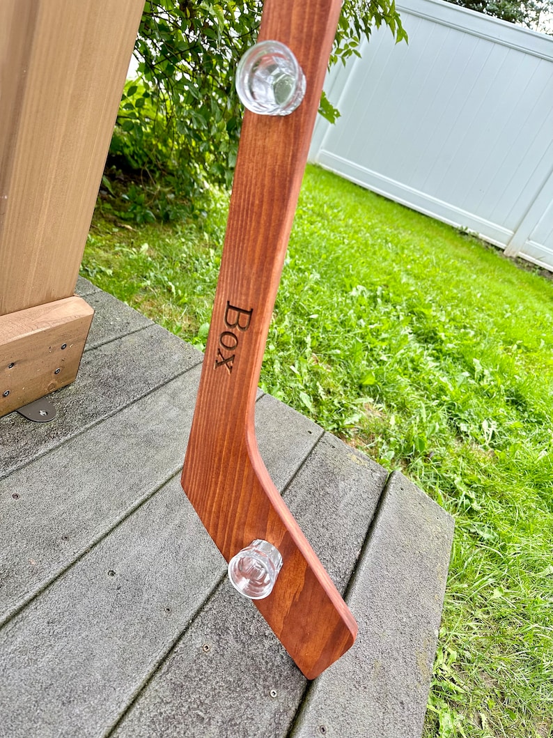 Puede incluir: Un palo de hockey de madera con dos vasos de chupito de vidrio transparente unidos a &eacute;l. El palo est&aacute; te&ntilde;ido de un marr&oacute;n rojizo oscuro y tiene la palabra "BOX" grabada en &eacute;l.
