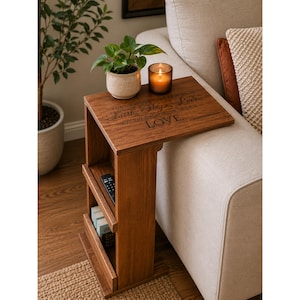 May include: A brown wooden C-shaped side table with a flat top surface featuring the engraved text Faith, Hope, Love, but the greatest of these is LOVE. The table includes two lower shelves holding a black remote control and small books.