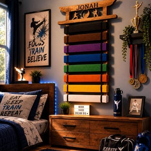 Op de afbeelding: Een houten wandrek voor vechtsportbanden met de naam JONAH bovenaan, met acht gekleurde banden in zwart, bruin, paars, blauw, groen, oranje, geel en wit, voorzien van silhouetten van vechtsporters.