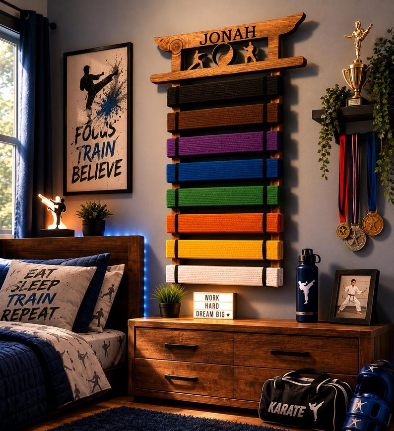 Op de afbeelding: Een houten wandrek voor vechtsportbanden met de naam JONAH bovenaan, met acht gekleurde banden in zwart, bruin, paars, blauw, groen, oranje, geel en wit, voorzien van silhouetten van vechtsporters.