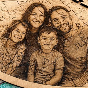 May include: A heart-shaped wooden jigsaw puzzle featuring a laser-engraved photograph of a family of four. The image depicts two adults and two children smiling, with the puzzle pieces clearly defined by dark, interlocking lines across the surface.