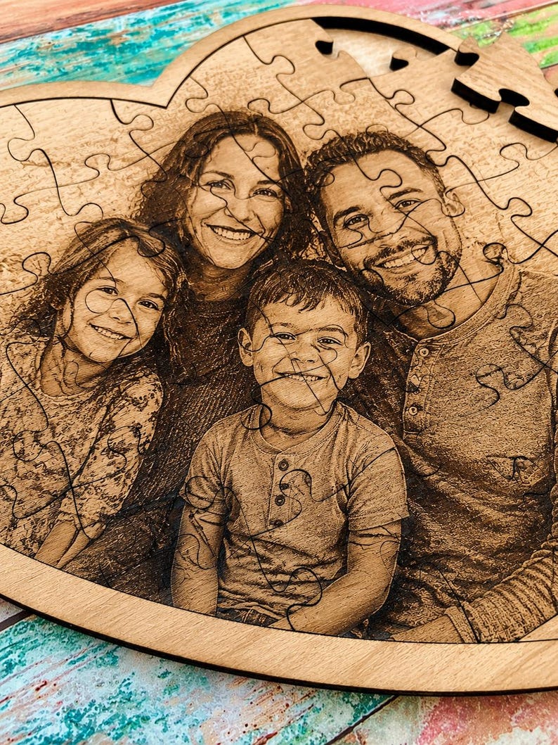 May include: A heart-shaped wooden jigsaw puzzle featuring a laser-engraved photograph of a family of four. The image depicts two adults and two children smiling, with the puzzle pieces clearly defined by dark, interlocking lines across the surface.
