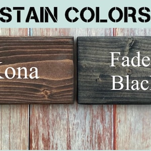 Op de afbeelding: Twee rechthoekige houten blokken tonen beitskleuren. Het linkerblok is donkerbruin met de tekst Kona, en het rechterblok is antracietgrijs met de tekst Faded Black. De koptekst luidt STAIN COLORS en er staat een cirkelvormig logo in de hoek.