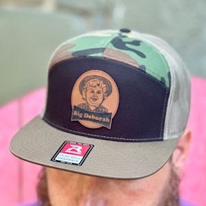 May include: A flat bill snapback hat with a camouflage top, black front panel, and a tan brim. The hat features a leather patch with the text "Big Deborah" and a cartoon image. The hat has a mesh back and a tag that reads "Flat Bill Snapback".