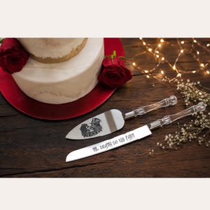 May include: A silver cake knife and server set with clear, faceted acrylic handles. The knife blade is engraved with the text "TIL DEATH DO US PART" and the server features an etched graphic of two skeletons embracing in top hats.