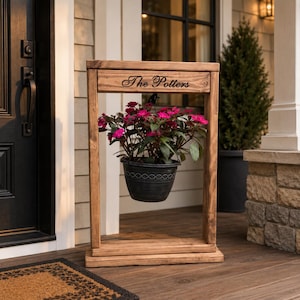 May include: A rectangular wooden stand with a natural brown finish featuring the text The Potters engraved on the top horizontal beam. A dark gray plastic planter containing a vibrant pink flowering plant hangs from the center of the frame.