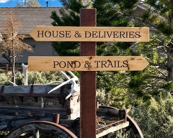 May include: A vertical dark brown wooden post featuring two horizontal light-colored wooden directional signs with pointed ends. The top sign reads HOUSE & DELIVERIES and the bottom sign reads POND & TRAILS in dark, engraved, capitalized lettering.