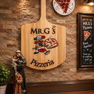 Może przedstawiać: Duży drewniany szyld ścienny w kształcie łopaty do pizzy z napisem MR.G'S Pizzeria i postacią z kresk&oacute;wki trzymającą pizzę. Powyżej znajduje się okrągły zegar z grafiką kawałka pizzy. Po prawej stronie tablica kredowa w ramie MR.G'S Pizzeria.