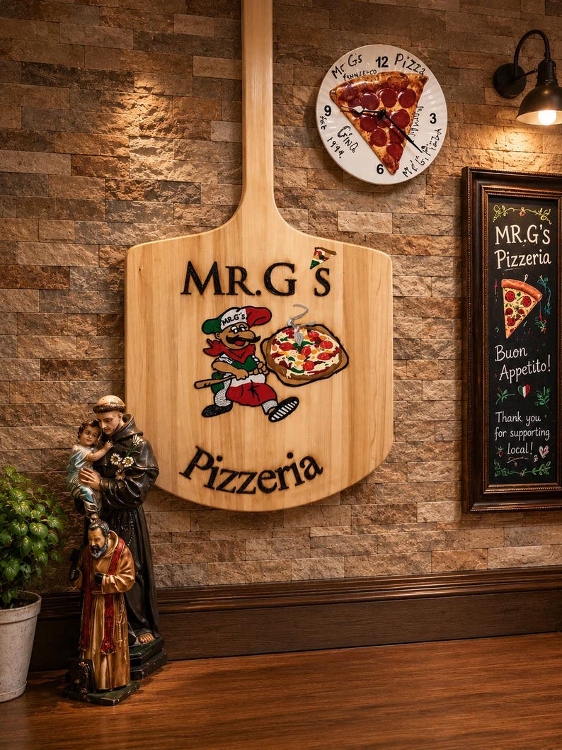 Może przedstawiać: Duży drewniany szyld ścienny w kształcie łopaty do pizzy z napisem MR.G'S Pizzeria i postacią z kresk&oacute;wki trzymającą pizzę. Powyżej znajduje się okrągły zegar z grafiką kawałka pizzy. Po prawej stronie tablica kredowa w ramie MR.G'S Pizzeria.