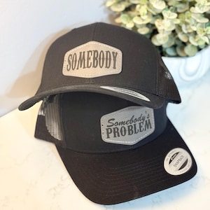 May include: Two black trucker hats with gray patches. The top hat has the word "SOMEBODY" on the patch. The bottom hat has the words "Somebody's PROBLEM" on the patch.