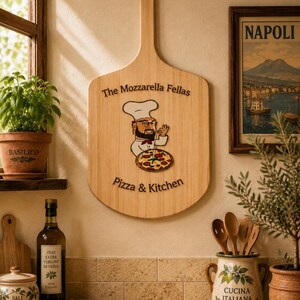 May include: A light-colored wooden pizza peel featuring a graphic of a chef with a beard and hat holding a pizza. The text reads The Mozzarella Fellas Pizza & Kitchen. The peel is mounted on a wall between a potted basil plant and a framed picture.